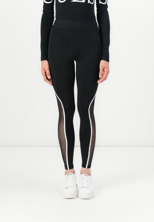 Black leggings with white side stripes, featuring sheer panels below the knee. Paired with white sneakers, highlighting a fitted design.