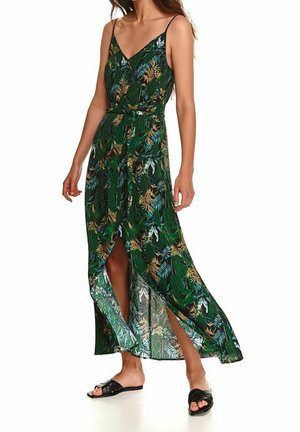 Nainen pukeutuneena vihreään tropiikkikuvioiseen, hihattomaan maksimekkoon, jossa on etuhiha ja mustat matalat liukusandaalit.