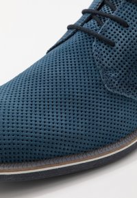 Chaussure en daim bleu perforé avec des lacets noirs, dotée d'une semelle en caoutchouc lisse et d'un subtil accent blanc le long de la base.