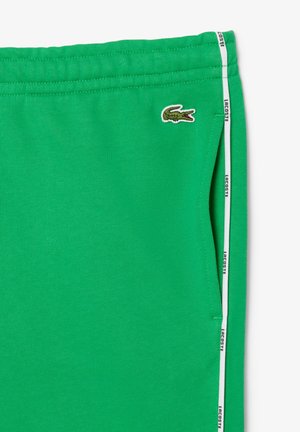 Grüne Jogginghose aus weichem Stoff, mit Seitentaschen, einem weißen Streifen mit "Lacoste"-Text und einem gestickten Krokodil-Logo.