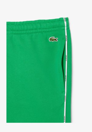 Calças de treino verdes feitas de um tecido macio, com bolsos laterais, uma tira branca com o texto "Lacoste" e um logótipo de crocodilo bordado.