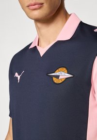 Tamsiai mėlynas polo marškinėlis su rožine apykakle, ant krūtinės – apvalus išsiuvinėtas Palermo FC logotipas ir rožinis Puma logotipas. Audinys su tekstūra.