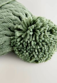 Chapeau en tricot vert avec un motif texturé et un grand pompon duveteux sur le dessus, mettant en valeur un matériau de fil doux et moelleux.