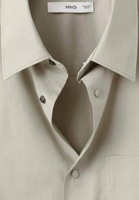 Beżowa koszula z kołnierzykiem i zatrzaskami, z widoczną metką marki "MNG" oraz szczegółami dotyczącymi rozmiaru wewnątrz dekoltu.