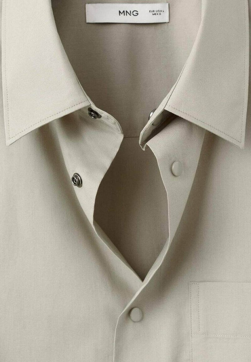Beige Hemd mit Kragen und Druckknöpfen, mit dem Markenschild "MNG" und Größenangaben im Halsbereich.