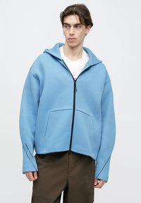 Φούτερ με φερμουάρ σε ανοιχτό γαλάζιο χρώμα, κατασκευασμένο από μαλακό ύφασμα, με κουκούλα, μπροστινή τσέπη και μαύρο φερμουάρ. Διαθέτει μακριά μανίκια με λεπτομερή ραφή.