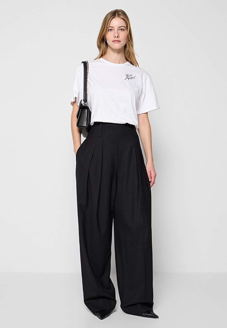 Weißes Baumwoll-T-Shirt mit aufgedrucktem Text, kombiniert mit hochgeschnittenen, weit geschnittenen schwarzen Hosen. Kleine Falten und schwarze spitze Schuhe runden den Look ab.