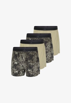 Athletischer dunkelhäutiger Mann steht barfuß, trägt beige Shorts mit schwarzem Bund und posiert mit einer Hand nahe dem Bund vor einfachem Hintergrund.