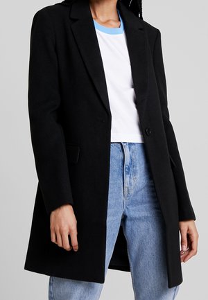 Sort enkeltknappet frakke båret over hvid T-shirt med lyseblå krave og lyseblå jeans, overkrop og øvre ben synlige, neutral baggrund.