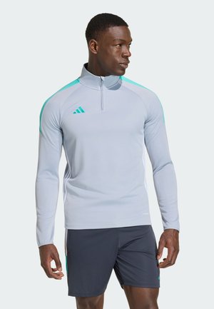Uomo atletico che indossa un top grigio chiaro a maniche lunghe con zip e dettagli turchesi e pantaloncini sportivi neri, guarda verso destra.