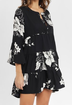 Woman wearing a black tiered dress with white floral print and three-quarter bell sleeves, standing against a plain light background.
