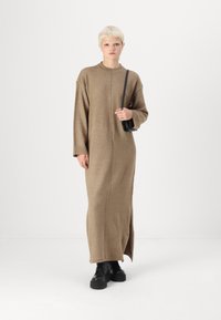 Robe longue en tricot marron avec un col montant, une fente sur le côté et des manches surdimensionnées, assortie à des bottines noires et un petit sac.