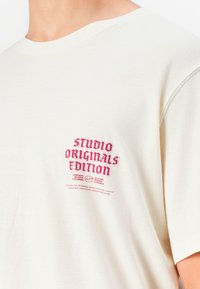 Bežio spalvos marškinėliai su raudonu užrašu "STUDIO ORIGINALS EDITION" ir mažesniu papildomu tekstu ant kairiojo krūtinės pusės, dėvimi asmens.
