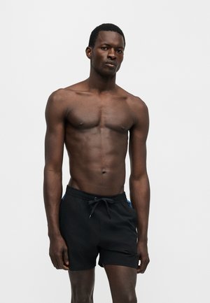 Homme à la peau foncée, torse nu, portant un short de bain noir, debout devant un fond clair uni, regardant devant lui.