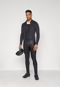 Tenue de cyclisme comprenant un maillot à manches longues noir ajusté, un gilet texturé, des leggings noirs et un casque noir, conçue pour la performance.