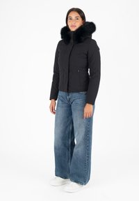Chaqueta acolchada negra con un gran cuello de piel sintética negra, bolsillos con cremallera y un diseño ajustado, combinada con unos vaqueros holgados azules y zapatillas blancas.