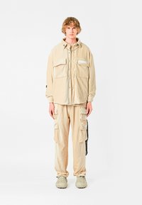 Conjunto de camisa y pantalones de pana beige con cuatro bolsillos delanteros, corte relajado y tela texturizada. Combinado con zapatillas texturizadas de color gris claro.