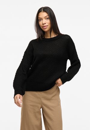 Maglione nero a maglia testurizzata con orlo e polsini a coste, caratterizzato da un classico colletto rotondo e una vestibilità leggermente ampia. Indossato con pantaloni beige.