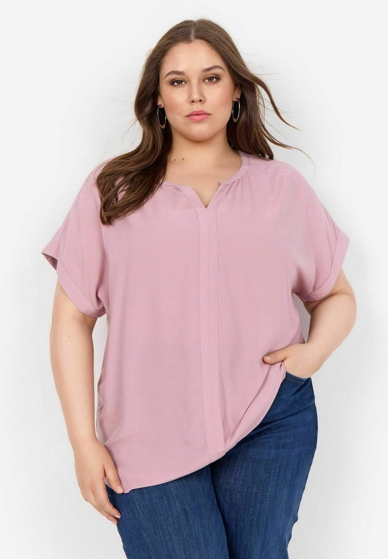 Blusa rosa chiaro a maniche corte con scollatura a V, caratterizzata da una cucitura centrale. Abbinata a jeans blu scuro, con texture liscia e vestibilità rilassata.