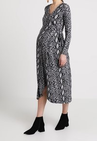 Kobieta w ciąży ubrana w długą, czarną i białą sukienkę w wężowy wzór z długimi rękawami, nosząca czarne botki za kostkę, stojąca na jednolitym tle.