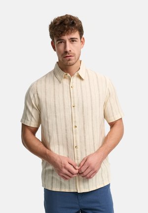 Mężczyzna o krótkich, kręconych włosach, ubrany w beżową, krótką koszulę na guziki z pionowymi paskami oraz niebieskie spodnie, stoi na neutralnym tle.