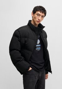 Schwarze Daunenjacke mit hohem Kragen, Reißverschluss und elastischen Bündchen. Verfügt über eine matte Textur und ein minimalistisches Design.