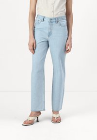 Svetlo modri jeans s ravnim krojem in sprednjimi žepi, kombinirani z belimi čevlji na peto z naramnicami. Tkani material ima mehak, nekoliko obrabljen videz.