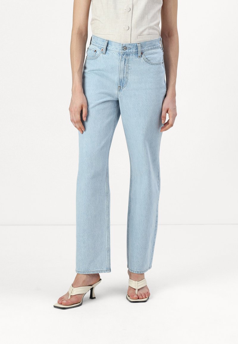 Svetlo modri jeans s ravnim krojem in sprednjimi žepi, kombinirani z belimi čevlji na peto z naramnicami. Tkani material ima mehak, nekoliko obrabljen videz.