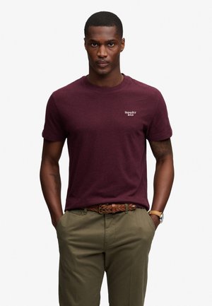 Hombre con camiseta Superdry granate, pantalones verde oliva, cinturón trenzado marrón y reloj de pulsera, de pie con las manos en los bolsillos.