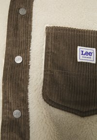 Brązowa kurtka z sztruksu z kremowym futerkiem, z metalowymi guzikami i kieszenią oznaczoną napisem "Lee WORKWEAR" z pionowymi prążkami.