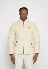 Veste en polaire couleur crème avec un col haut, fermeture éclair à l'avant et poches latérales. Présente un petit logo de la marque sur la poitrine et une surface texturée.