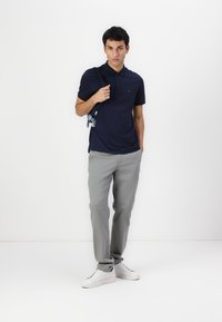 Námornícky polo tričko, krátke rukávy, golier, hladká textúra. Svetlošedé nohavice so striha rovného strihu. Biele tenisky. Model drží popruh tašky.