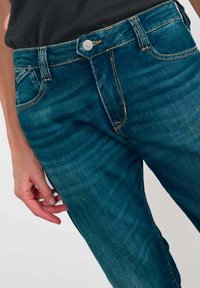 Jean en denim bleu avec une légère décoloration, taille mi-haute, design à cinq poches, et surpiqûres orange contrastées. Détail de fermeture à bouton en argent.