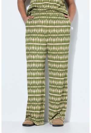 Person, die grüne und beige gemusterte, weit geschnittene Hosen mit den Händen in den Taschen trägt, steht auf weißem Boden vor weißer Wand.