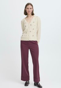 Cardigan color crema con ricamo floreale, scollo a v, e chiusura con bottoni abbinato a pantaloni larghi bordeaux con risvolti.