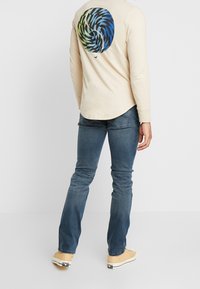 Man med beige långärmad skjorta med blå och grön virveldesign på ryggen, blå jeans och beige sneakers, som står mot en vit bakgrund.