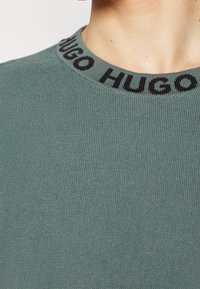 Gros plan sur le col d'un pull tricoté vert avec le texte noir « HUGO » autour de l'encolure sur la partie supérieure de la poitrine et du cou d'une personne.