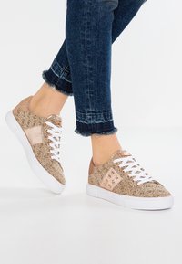 Béžové tenisky s vzorem, bílými tkaničkami a gumovými podrážkami, s texturovanou tkaninou a kovovými akcenty, v kombinaci s krátkými modrými džínovými kalhotami.