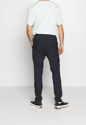 Man draagt een wit T-shirt, donkere geruite slim-fit broek en zwarte lage sneakers, staat tegen een witte achtergrond, achteraanzicht.