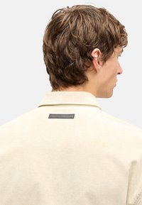 Koszula z kołnierzykiem z jasnobeżowego sztruksu, o subtelnej fakturze, z ciemną metką z tyłu przy szyi.