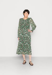 Vestido longo floral com um padrão verde, preto e branco. Apresenta mangas longas transparentes e um decote franzido, caindo abaixo do joelho.