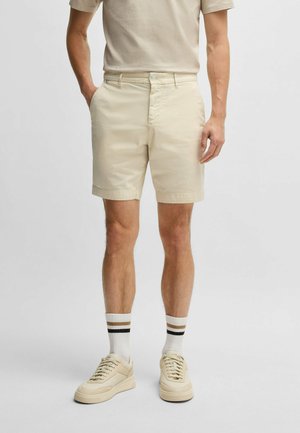 Beige katoenen shorts met een vlakke voorkant, zijzakken en een knoopsluiting, gecombineerd met crèmekleurige sneakers en witte sokken met bruine strepen.