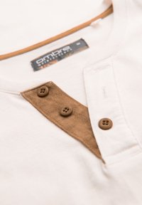 Koszulka henley z krótkim rękawem w kolorze kremowym, z brązowym zapięciem z zamszu i trzema brązowymi guzikami. Metka widoczna przy dekolcie. Gładka faktura materiału.