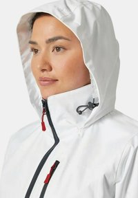 Chaqueta impermeable blanca con capucha ajustada, que presenta cremalleras negras y tiradores rojos. La vista en primer plano muestra la textura de la tela y los detalles del diseño.
