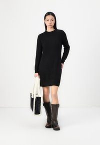 Vestido de punto negro con mangas largas; combinado con botas de cuero marrón hasta la rodilla y un bolso de pelo con ribete color crema.