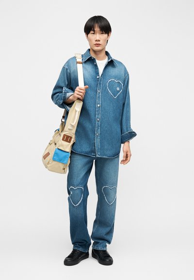 Άτομο που φοράει τζιν πουκάμισο και τζιν με σκισμένες καρδιές, μαύρα παπούτσια, λευκό πουκάμισο και μπεζ τσάντα ώμου με μπλε τσέπη.