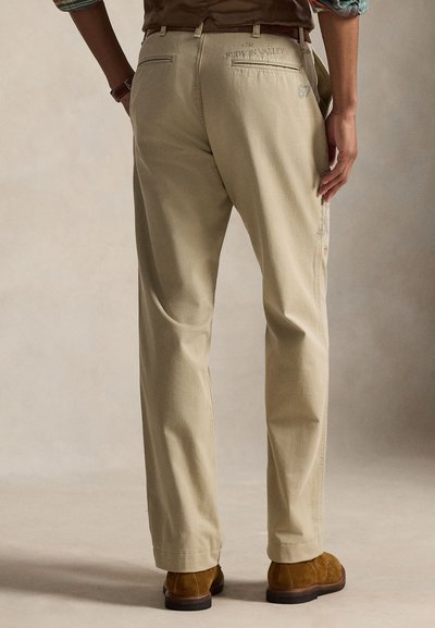 Beige bomullsbukser med rett snitt, med baklommer og brodert tekst. Stoffet har en glatt tekstur. Egnet for casual bruk.