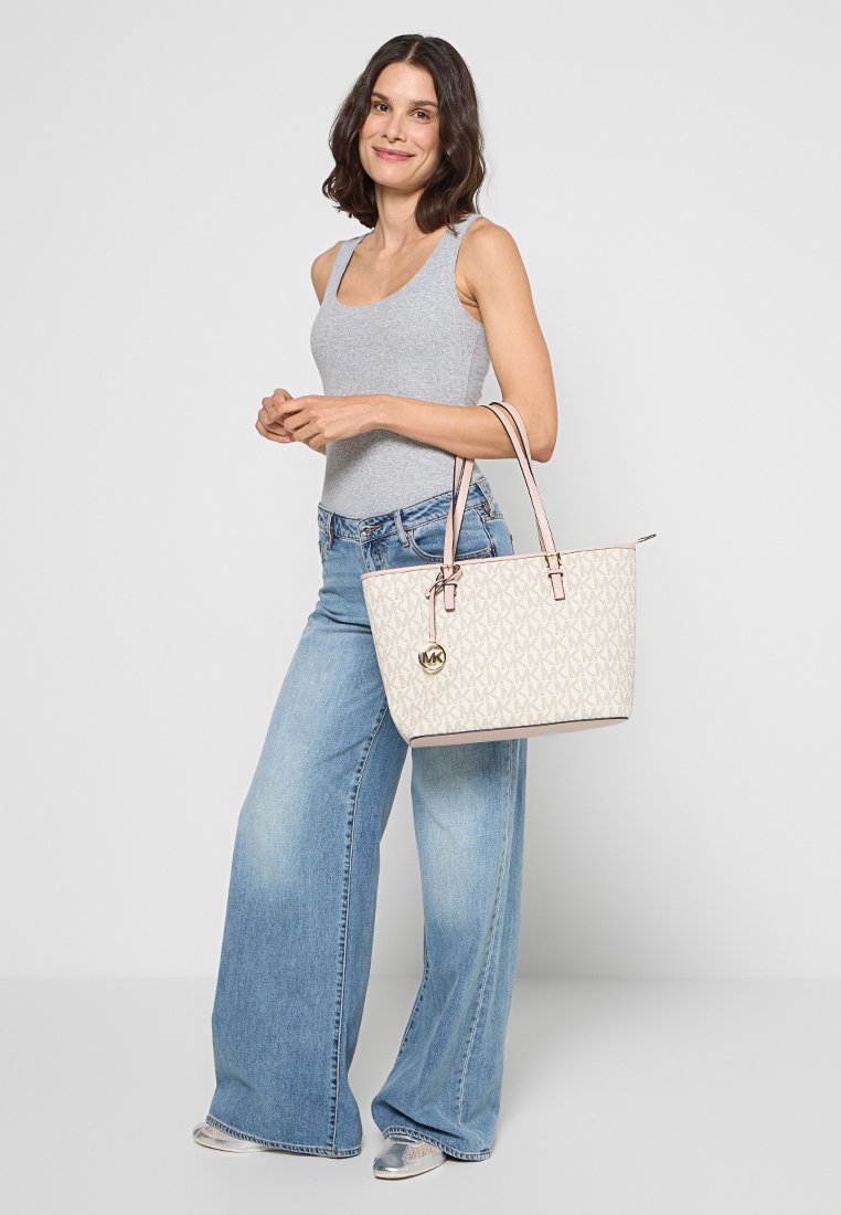 Beige Shopper Tasche mit MK-Logo, aus synthetischem Material, rosa Akzenten, strukturiertem Design und zwei stabilen Griffen. Modell trägt helle Jeans und ein graues Tanktop.