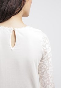 Top bianco con intricate maniche lunghe in pizzo. Presenta una chiusura a chiave sul retro e un tessuto leggermente strutturato, che evidenzia dettagliati motivi floreali.