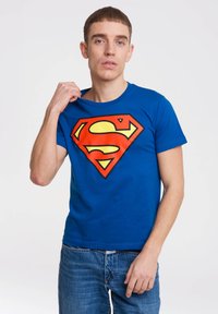 Young man wearing blue t-shirt with large red and yellow Superman logo, pulling at the neckline, standing against white background.